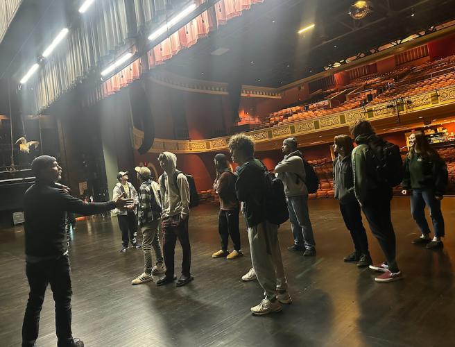 A group of students stands on a stage with their backs to the empty theater seats