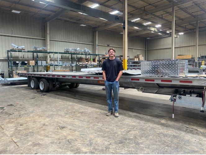 A man stands in a warehouse in front of a flatbed truck, wearing safety glasses