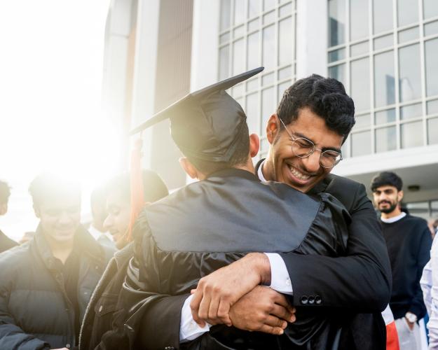 Two men, one wearing a graduation cap and gown, embrace as the sun shines behind them