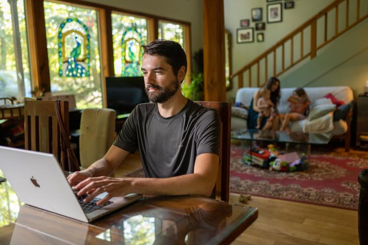 Online student on computer in livingroom with fmaily in the background