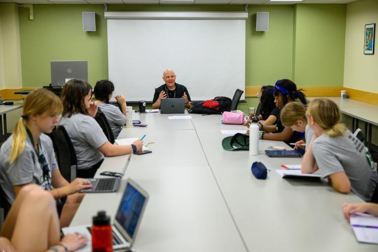 High school students sit around a table in a classroom on the Athens campus listening attentively to a workshop presenter at the 2024 High School Journalism Workshop at Ohio University.