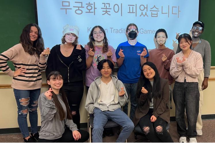 A group poses in front of a classroom, smiling and making a gesture with their hands