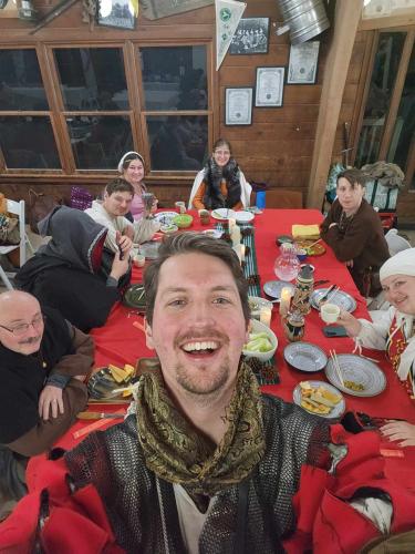 A man dressed in Medieval garb takes a selfie as a similarly dressed group sits behind him at a dining table