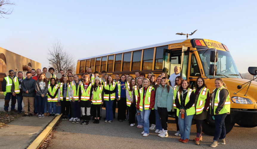 High school students from the region are show wearing safety vests while standing outside of a school bus