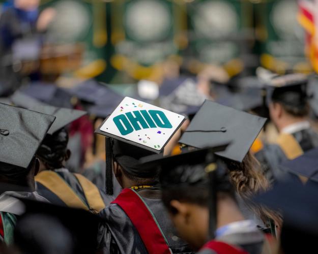 An OHIO student wears a commencement cap that says "OHIO" while sitting in a large group of students during the Commencement ceremony in the Convocation Center