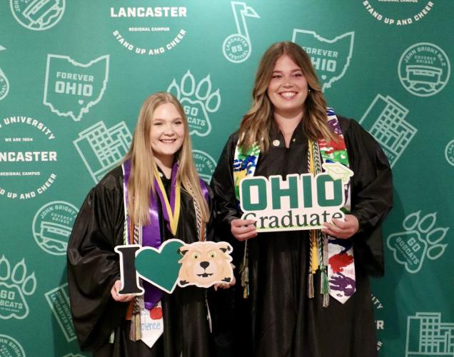 Two OHIO Lancaster graduates are shown in their graduation robes while holding OHIO Graduate signs