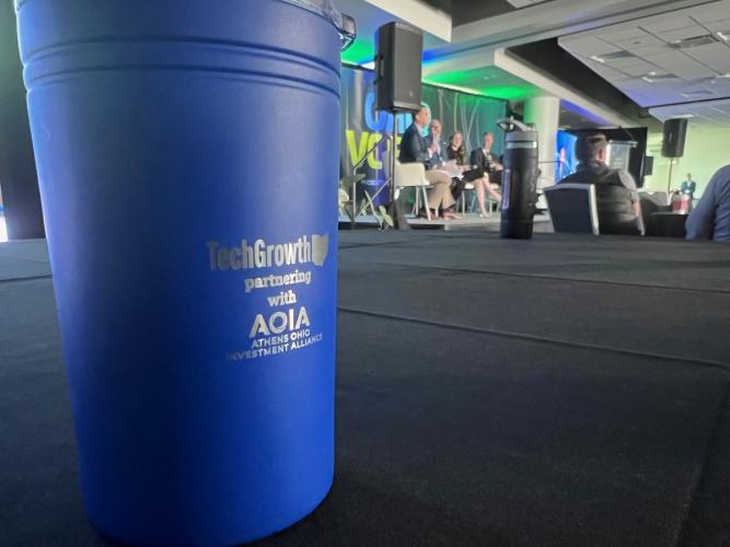 A sign that says TechGrowth partnering with AOIA Athens OHIO Investment Alliance is shown at a media event where speakers are sitting on a stage