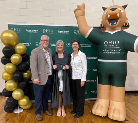 Mary Stephan is shown in commencement robe, holding her cap, standing with Scott Titsworth and Ann Bainbridge while they stand next to balloons and a blow-up figure of Rufus wearing a Scripps College of Communication shirt