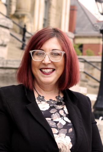 Female, smiling with shoulder length red hair, wearing glass, black and white blouse with a black blazer