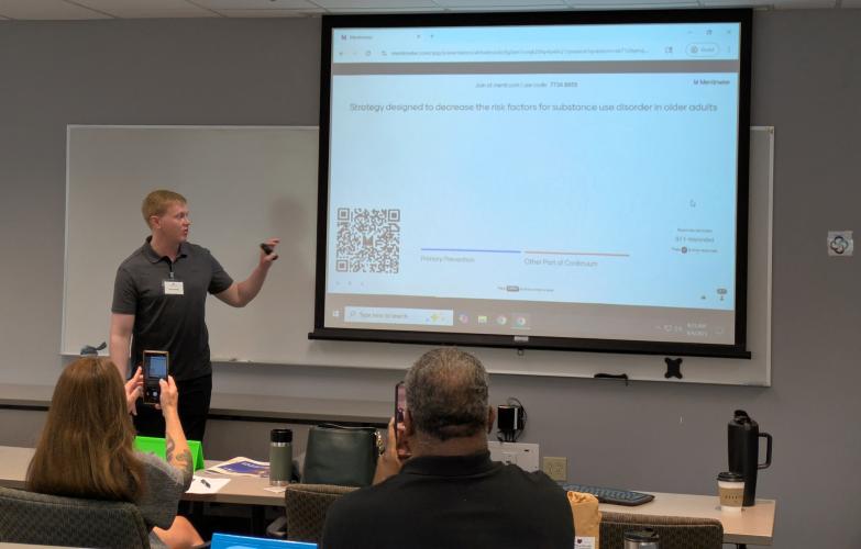 An instructor from the Ohio Center of Excellence for Behavioral Health Prevention  and Promotion points  at a power point screen while leading a class presentation