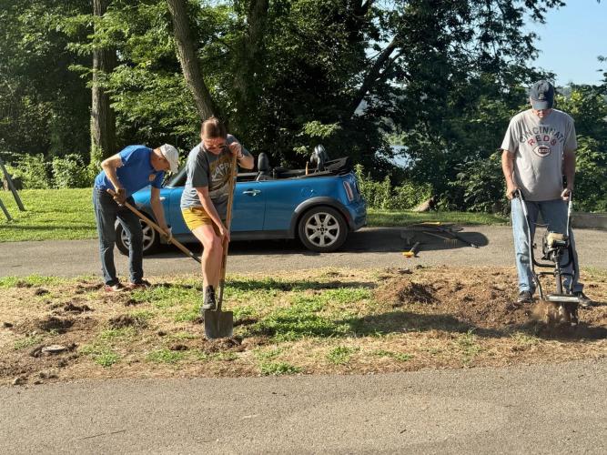 Three people work on outdoor garden space with shovels, rakes, tiller.