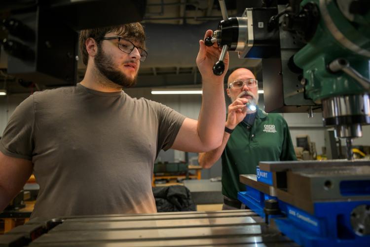 An OHIO student works on manufacturing equipment at the Workforce Development Center while an OHIO faculty member stands nearby and shines a flashlight on the equipment.