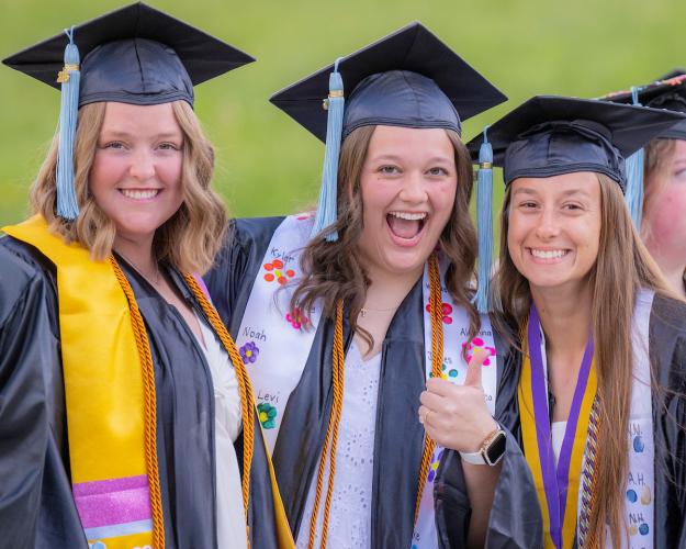 Three OHIO graduates smile together while wearing their caps and gowns, standing outside after the Graduation Recognition Ceremony at Ohio University Lancaster