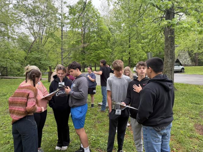 Students stand outdoors in small groups with clip boards