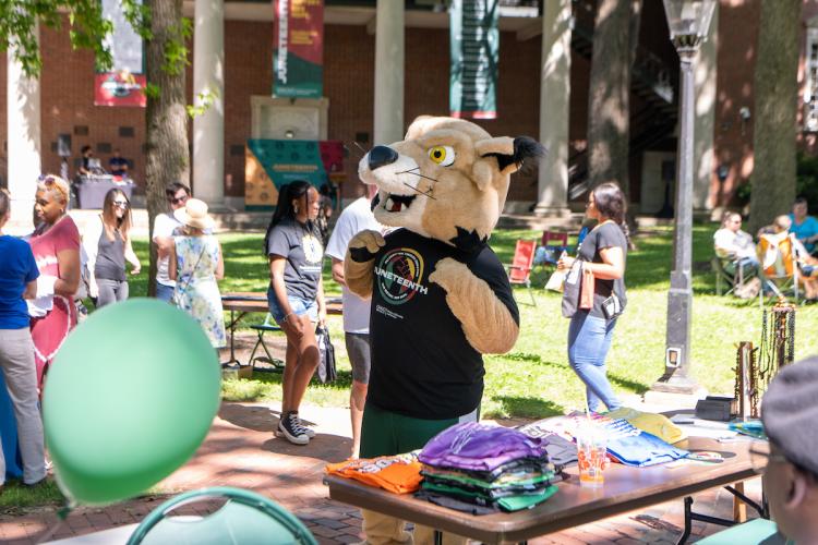 The Ohio University Rufus mascot wears a Juneteenth shirt and walks near a table at the Juneteenth Festival