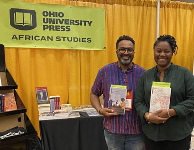 Jody Benjamin and Ademide Adelusi-Adeluyi hold their books and stand by a banner that says "Ohio University Press - African Studies"
