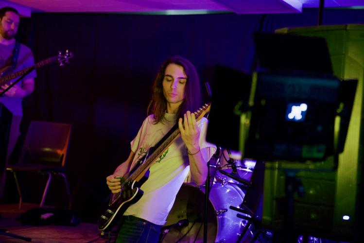 An individual plays guitar while another person plays a guitar in the background