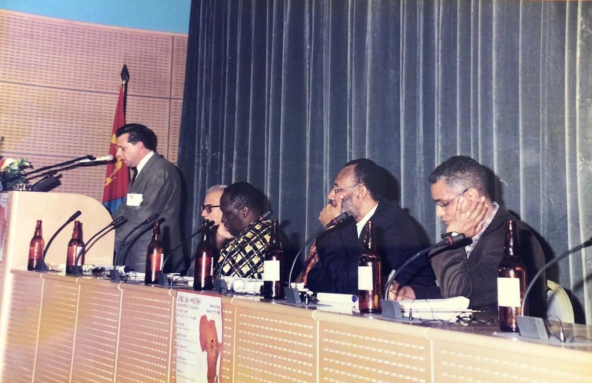 Standing at a podium, Ghirmai Negash speaks as Abdellatif Abdalla, Sheriff Hatata, Charles Cantalupo are seated and listening.