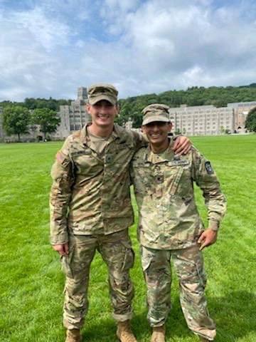 Two cadets stand side by side with arms wrapped around their shoulders in a field, smiling at the camera