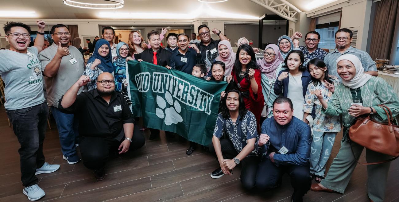 A large group of alumni and friends are shown holding an OHIO flag at an alumni event in Malaysia