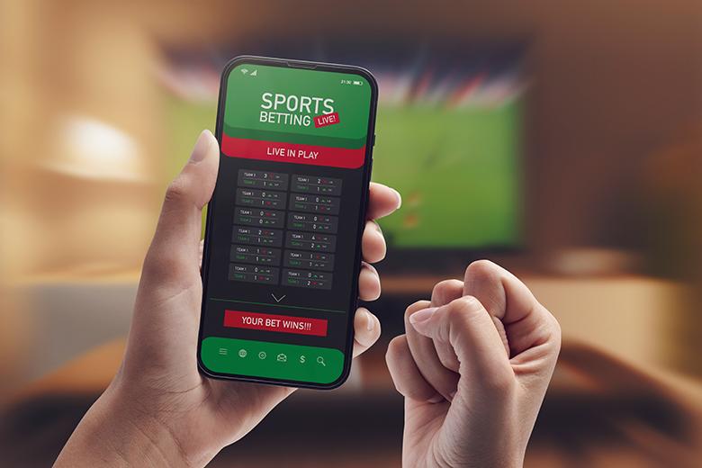 A sports bettor holds a phone with a sports betting app as they watch a game on television in the background.