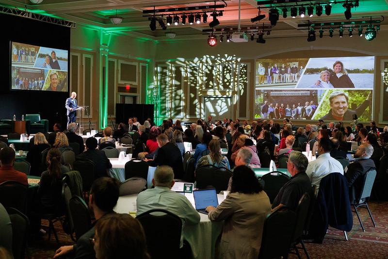 A speaker gives a presentation to a large crowd seated at tables in the Baker Center Ballroom