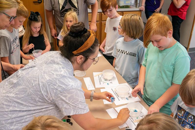 A teacher works with small jars of paint while students stand around her at a desk filled with bowls and paper