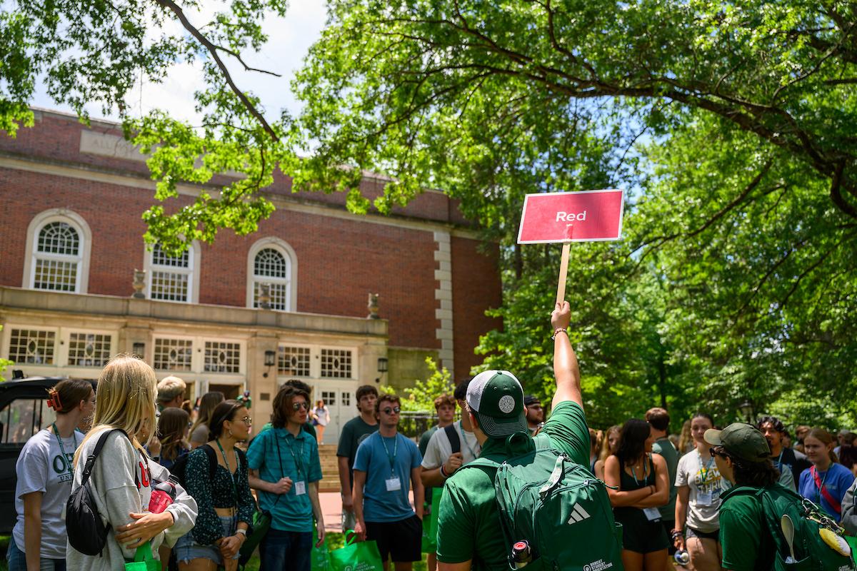An orientation leader holds up a sign that says "Red" for a group of students and family members standing outside on the College Green while taking part in Bobcat Student Orientation