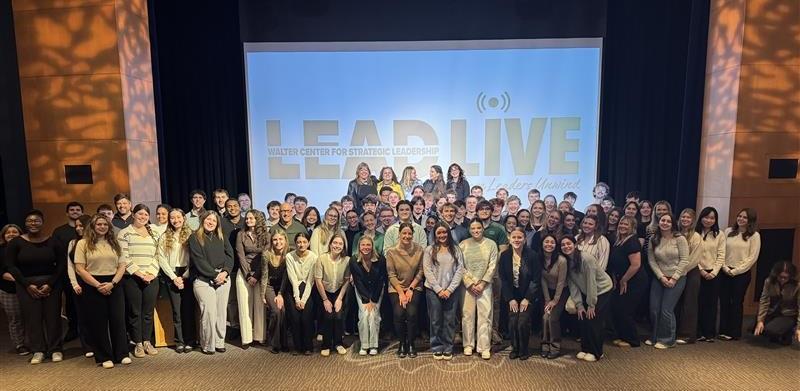 A large group of audience members pose with the speakers in front of the stage at the Lead Live event.
