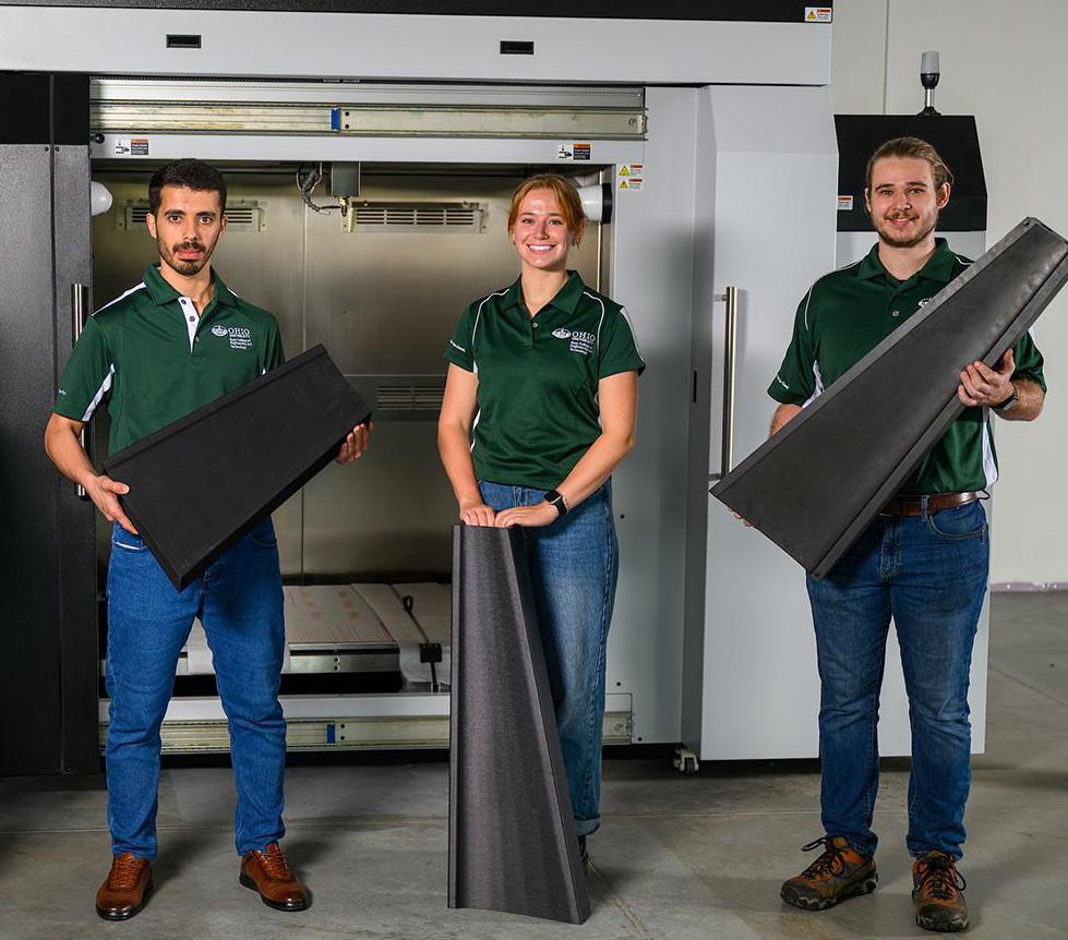 graduate students—Obieda Altarawneh (left), Grace Baranack, and Caleb Gula—holding 3D-printed carbon-enhanced composites fabricated using our large-scale FGF printer, shown in the background. 