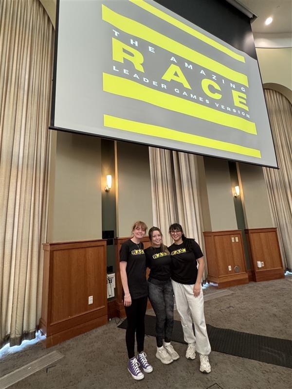 The members of the winning team, Morgan Elliott, Carolyn Bromley and Carsen Morin, stand below a graphic that says "The Amazing Race Leader Games Version."