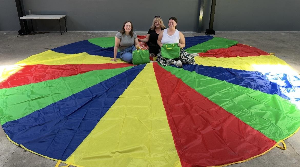 Three Ohio University representative sit on a colorful parachute that is flat in a circle on the floor