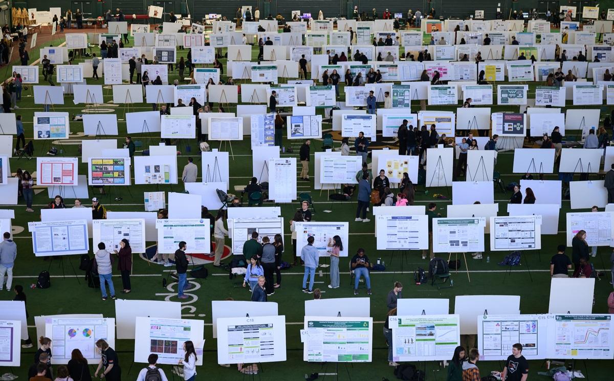 Rows of research posters, with students standing near them, are shown in the Walter Fieldhouse for the Student Research Expo.