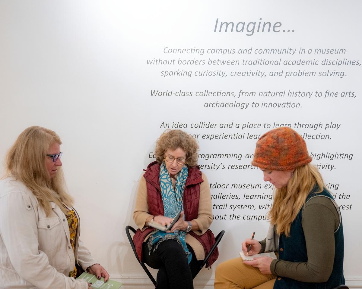 OHIO faculty and students sit in front of a wall that says "Imagine....Connecting campus and community in a museum without borders between traditional academic disciplines, sparking curiosity, creativity, and problem solving. World-class collection, from natural history to fine arts, archaeology to innovation." The wall has additional words, but the people are sitting in front of them.