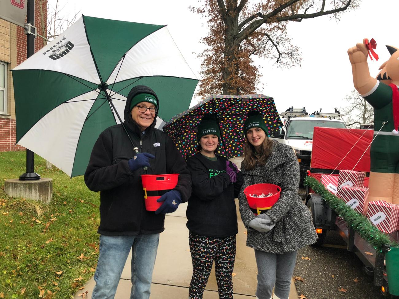 Male standing aside two female students dressed in hats and coats holding umbrellas and smiling
