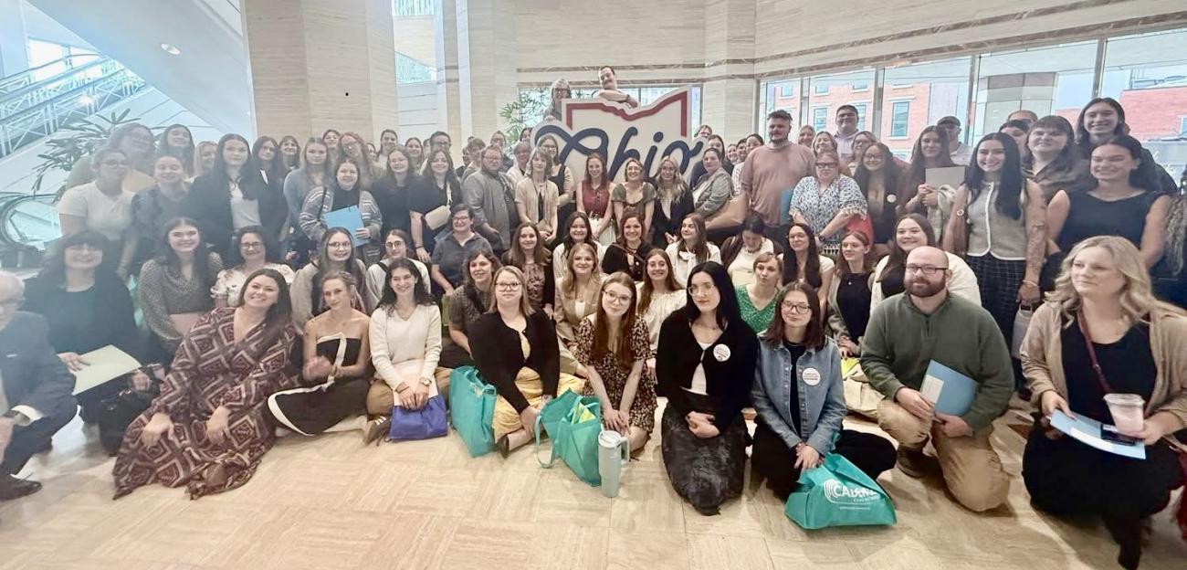 A very large group of OHIO students and faculty stand and sit together for a photo in front of an Ohio sign at the Ohio Statehouse.