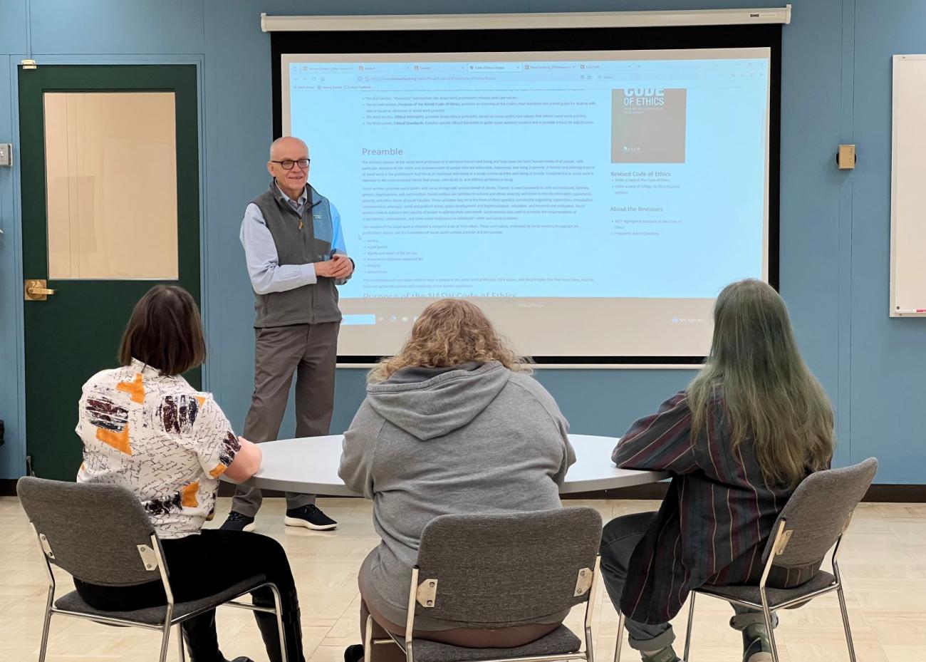 Older male standing next to a projection screen, lecturing to 3 students seated at a circular desk facing the instructor.