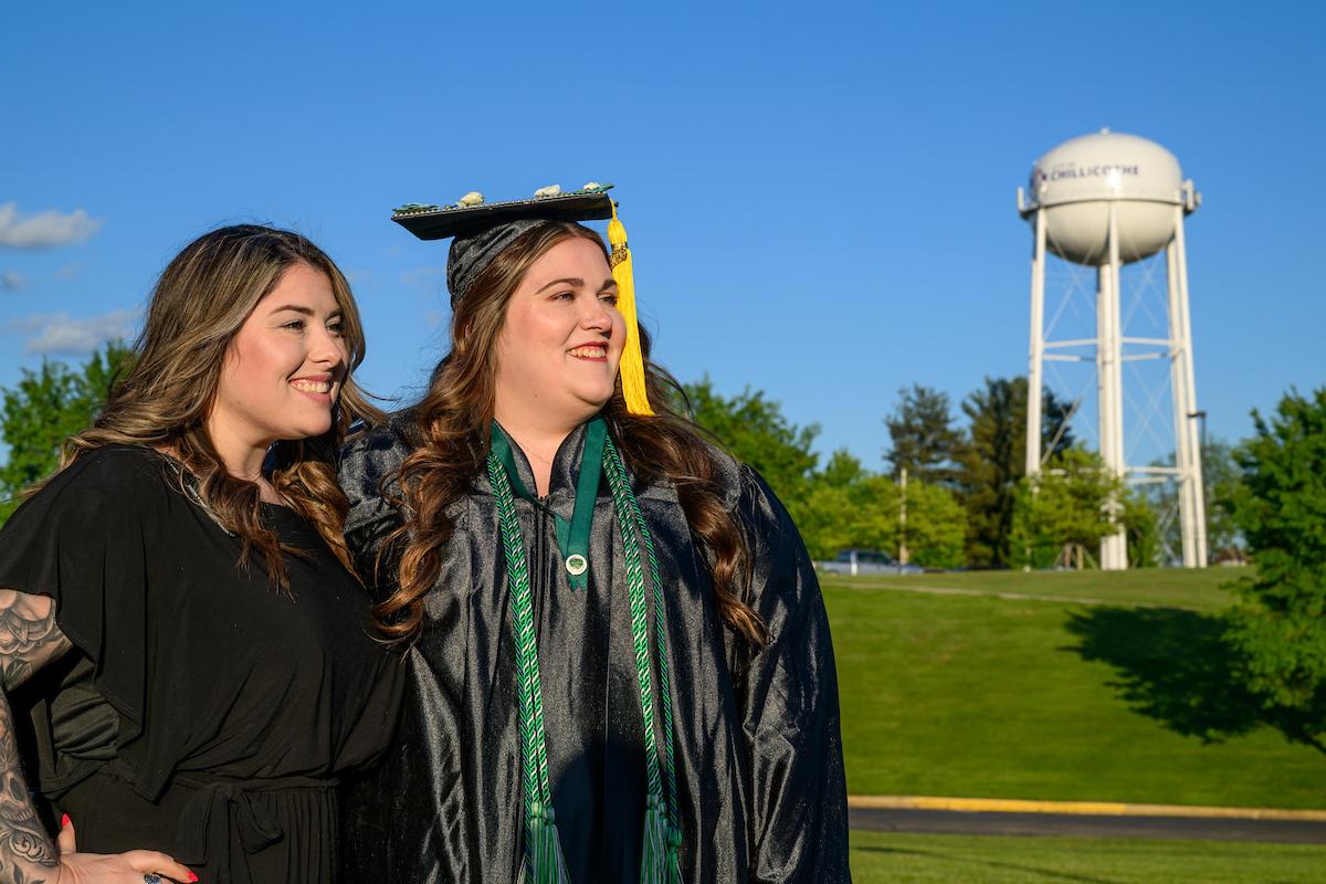 An OHIO graduate wearing the graduation cap and gown stands with a family member or friend outside while a Chillicothe water tower is shown in front of blue skies in the background