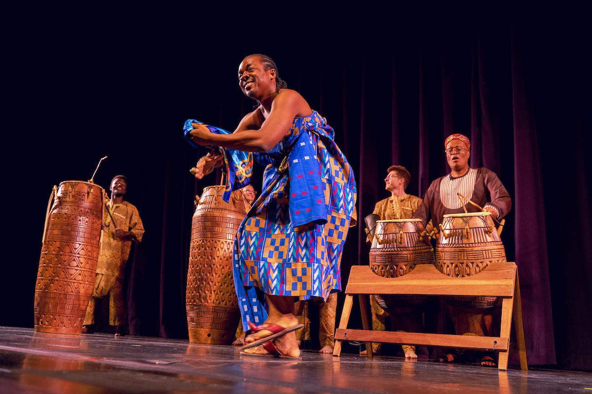 A performer dances on stage while drummers perform behind her. Dr.  Paschal Yao Younge is one of the drummers