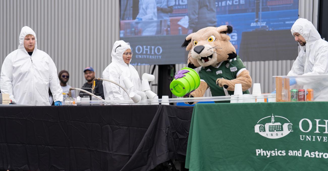 OHIO representatives in lab coats and hoods and the Rufus mascot perform a science demonstration at an Ohio University table at the Science Day event