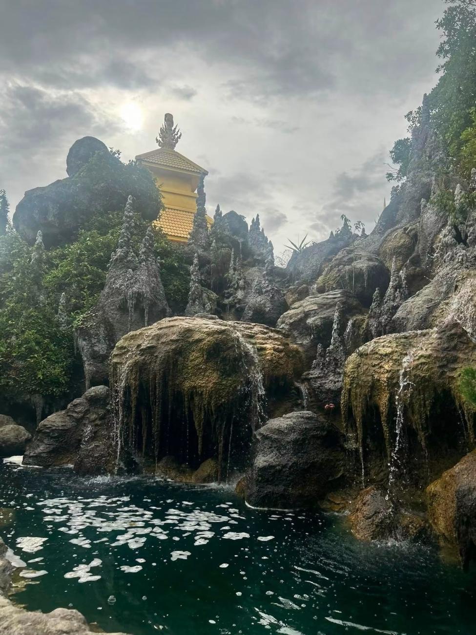 An image  from Thailand of water flowing over large  rocks to a pool below,  and a building is shown on land above the rocks
