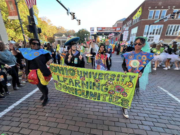 The Honey for the Heart  Parade marches down Court street while two people in the parade hold  a banner that says "Ohio University Learning Communities"