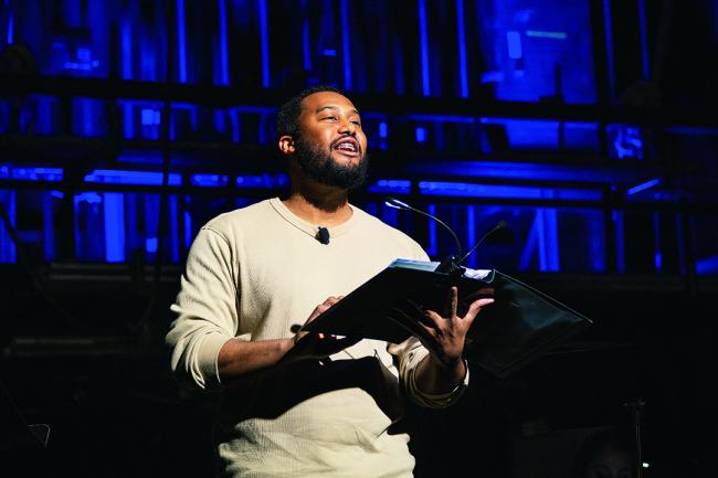 Black man with short hair, a beard, braces and a tan shirt stands in front of a microphone and a blue backdrop, reading from a black three-ring binder