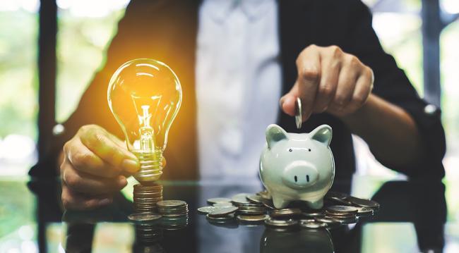 A person sorts coins into a piggy bank next to a glowing lightbulb