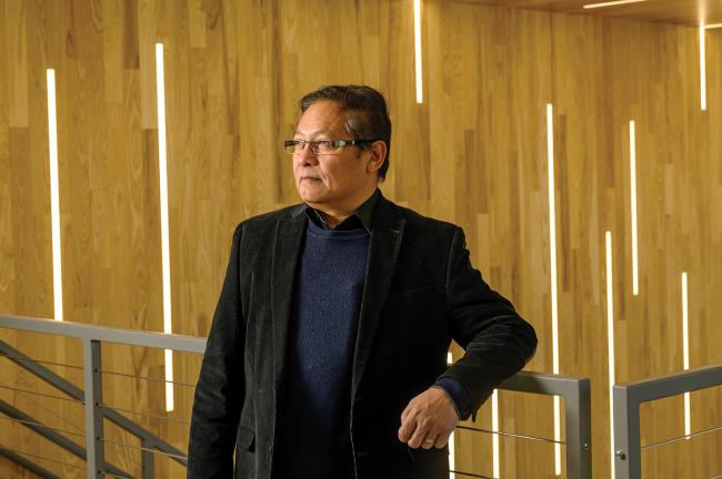 southeast Asian scientist Saw Wai Hla leans on a railing in front of a warm wood backdrop; he has dark hear and wears a black sport coat and glasses