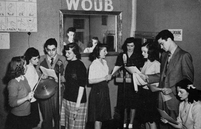 black-and-white photo of a group of students crowded around two microphones holding scripts in an early WOUB studio