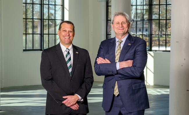  two white middle-age men pose, smiling, in suits in a large industrial-style room