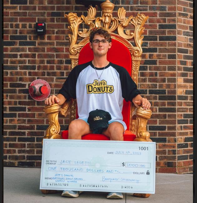 Jack Legath wears a Jeff's Donuts shirt and hat and sits on a throne after winning an eating competition