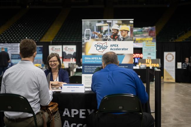 Three individuals talk at an APEX Accelerator table at an event