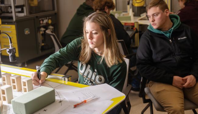 a female student in an Ohio University t-shirt creates a manufacturing model at a desk